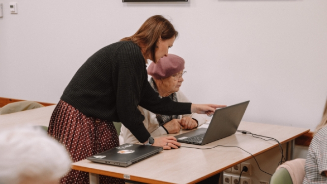 Sieviete palīdz seniorei pie klēpjdatora, norādot uz ekrānu izglītojošā nodarbībā