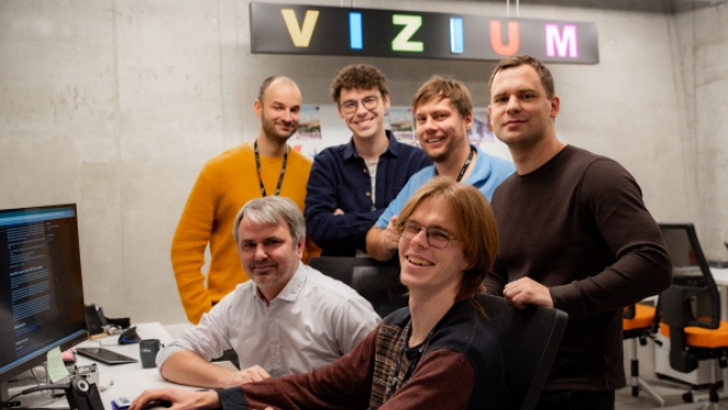 Sešu vīriešu grupa smaida kopbildei pie biroja galdiem, fonā redzams izgaismots uzraksts "VIZIUM".