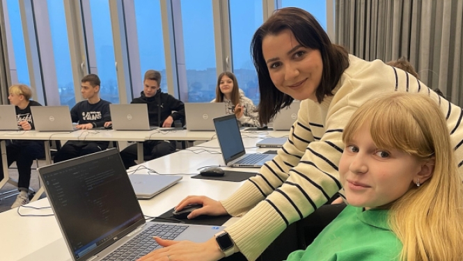 Sieviete svītrainā džemperī palīdz meitenei zaļā hūdijā programmēšanas nodarbībā pie klēpjdatora