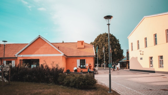 Skats uz pagalmu starp divām košām ēkām — oranžu un dzeltenu — saulainā dienā ar cilvēkiem fonā.