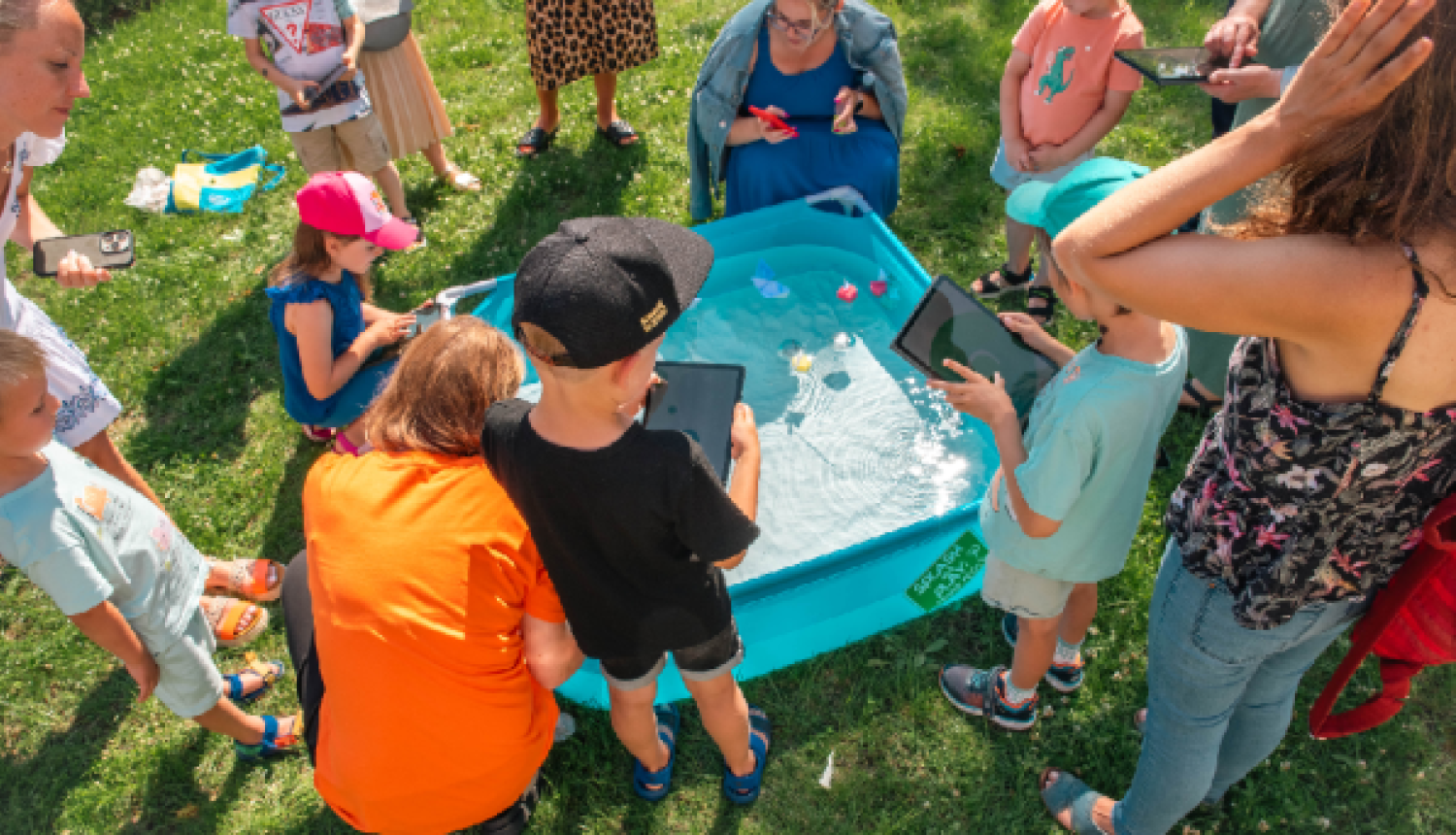 Bērni un pieaugušie brīvā dabā apkārt nelielam baseinam lieto planšetdatorus un viedtālruņus