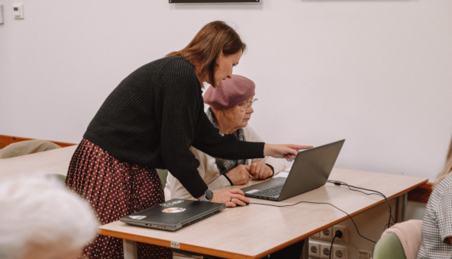 Sieviete palīdz seniorei pie klēpjdatora, norādot uz ekrānu izglītojošā nodarbībā