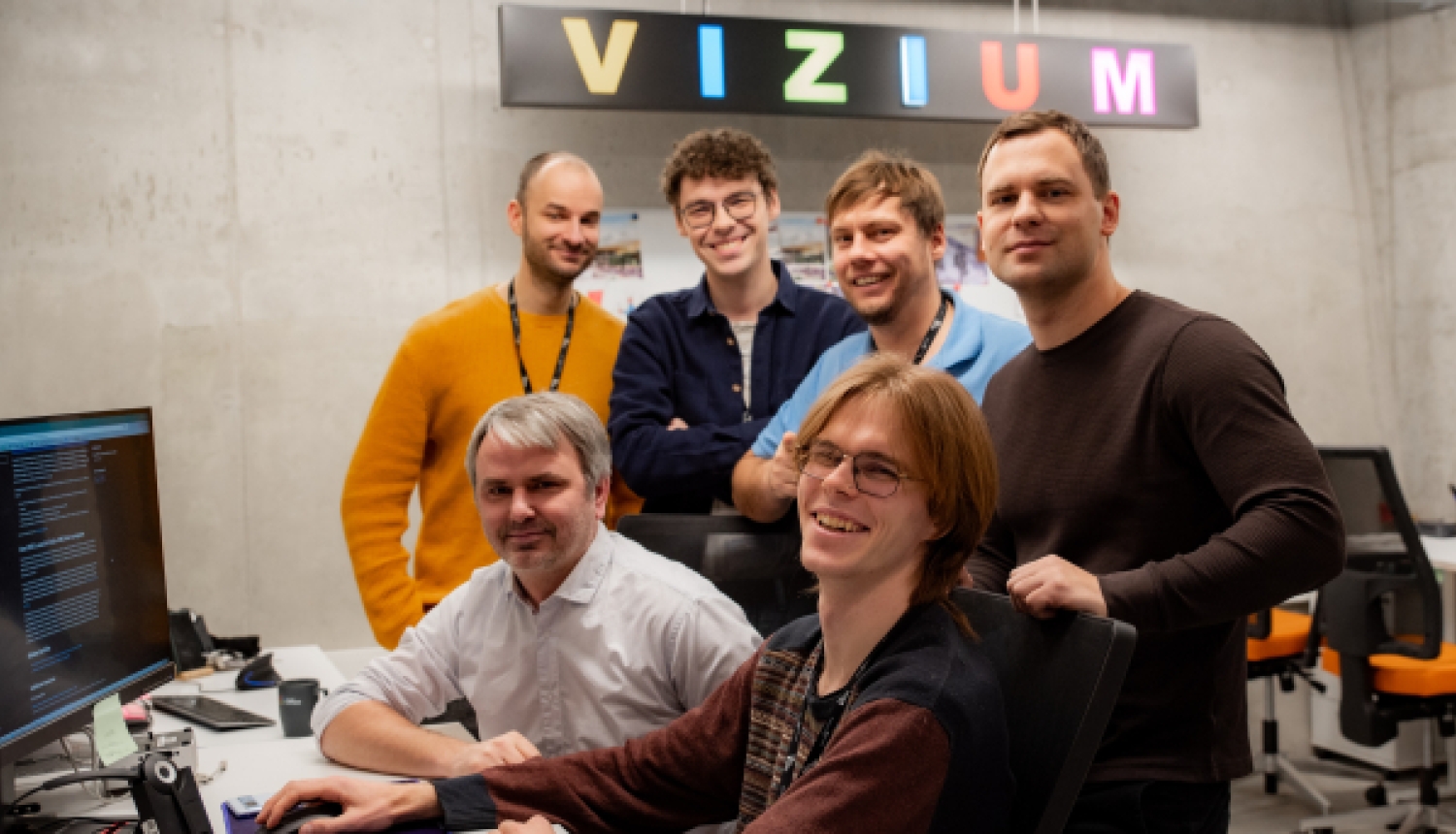 Sešu vīriešu grupa smaida kopbildei pie biroja galdiem, fonā redzams izgaismots uzraksts "VIZIUM".