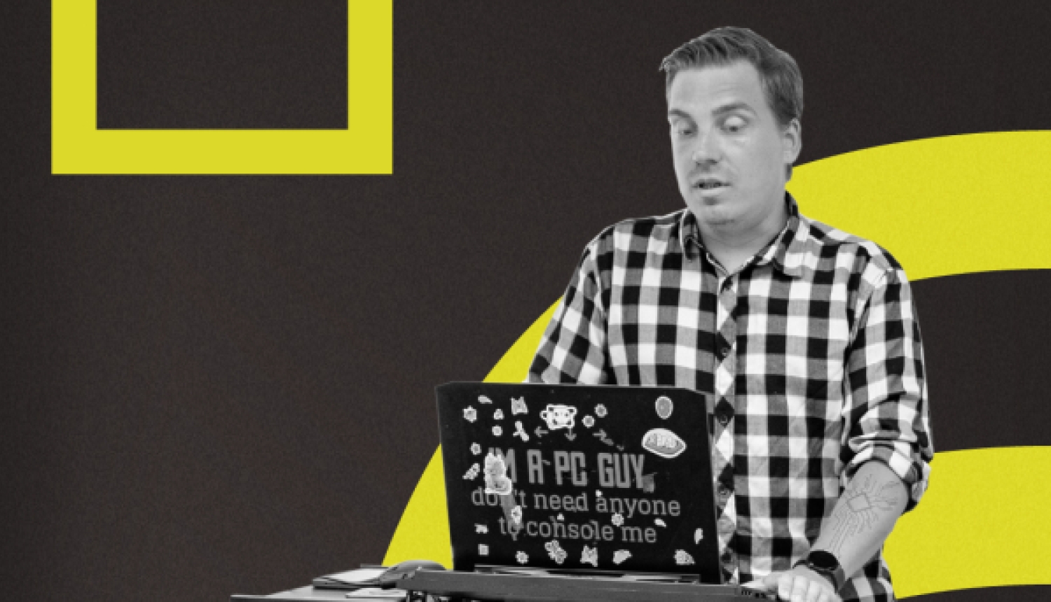 Vīrietis rūtainā kreklā stāv pie augstā galda un strādā ar klēpjdatoru, kura vāks ir noklāts ar uzlīmēm, uz tumša fona ar dzelteniem grafiskiem elementiem.