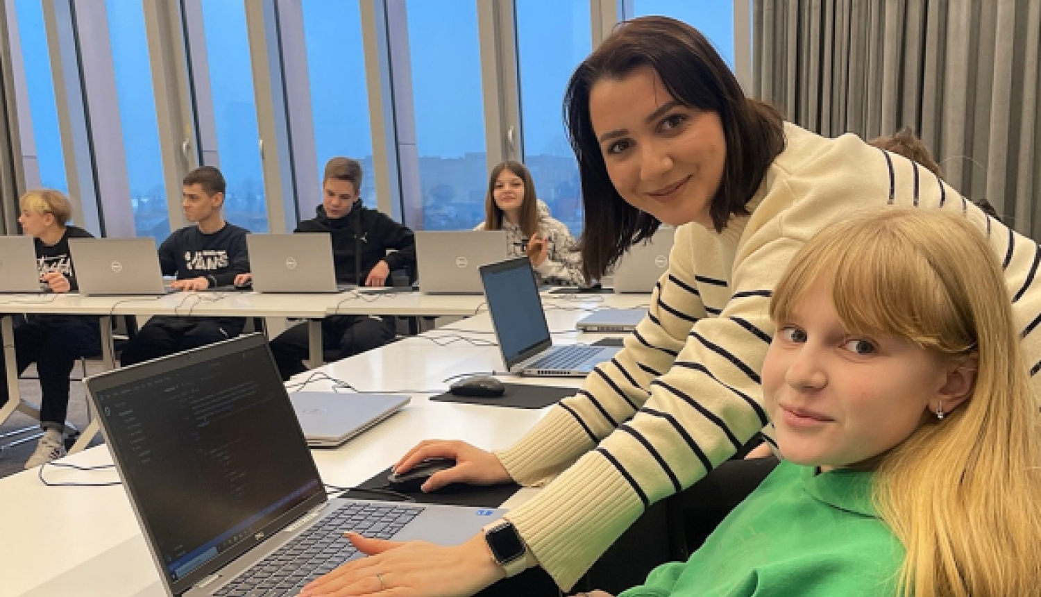 Sieviete svītrainā džemperī palīdz meitenei zaļā hūdijā programmēšanas nodarbībā pie klēpjdatora