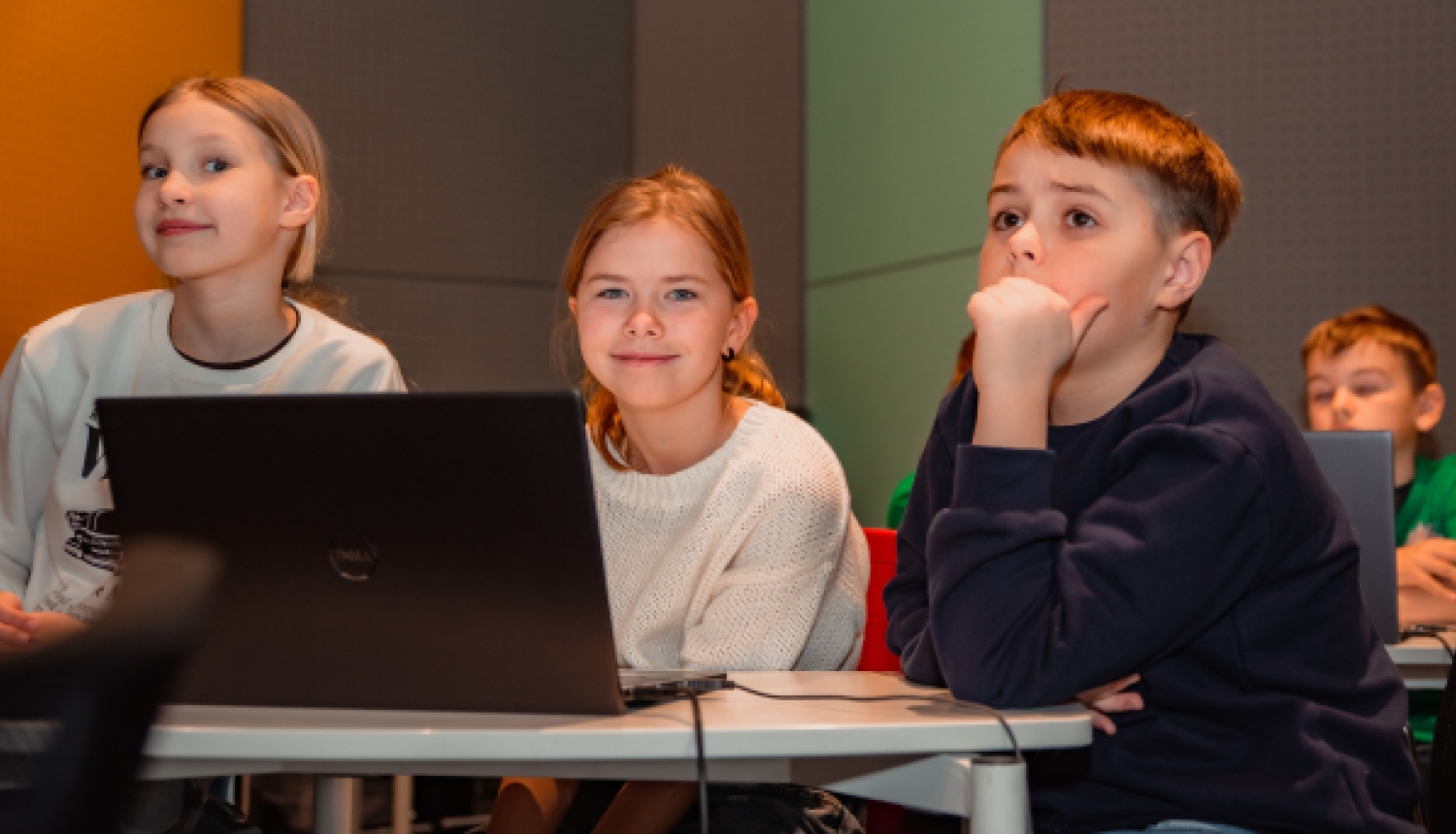 Trīs bērni sēž pie galda ar klēpjdatoru un uzmanīgi klausās nodarbību.