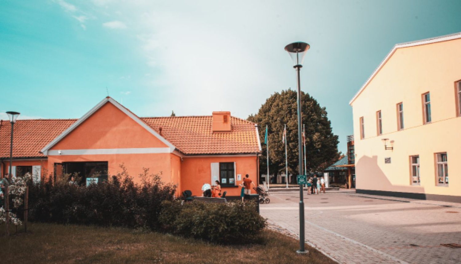 Skats uz pagalmu starp divām košām ēkām — oranžu un dzeltenu — saulainā dienā ar cilvēkiem fonā.