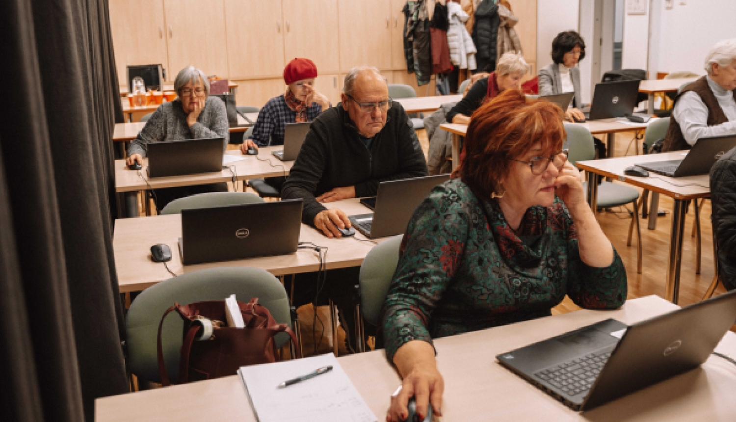 Vairāki seniori sēž pie galdiem mācību telpā un apgūst prasmes darbā ar klēpjdatoriem.
