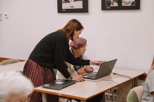 Sieviete palīdz seniorei pie klēpjdatora, norādot uz ekrānu izglītojošā nodarbībā