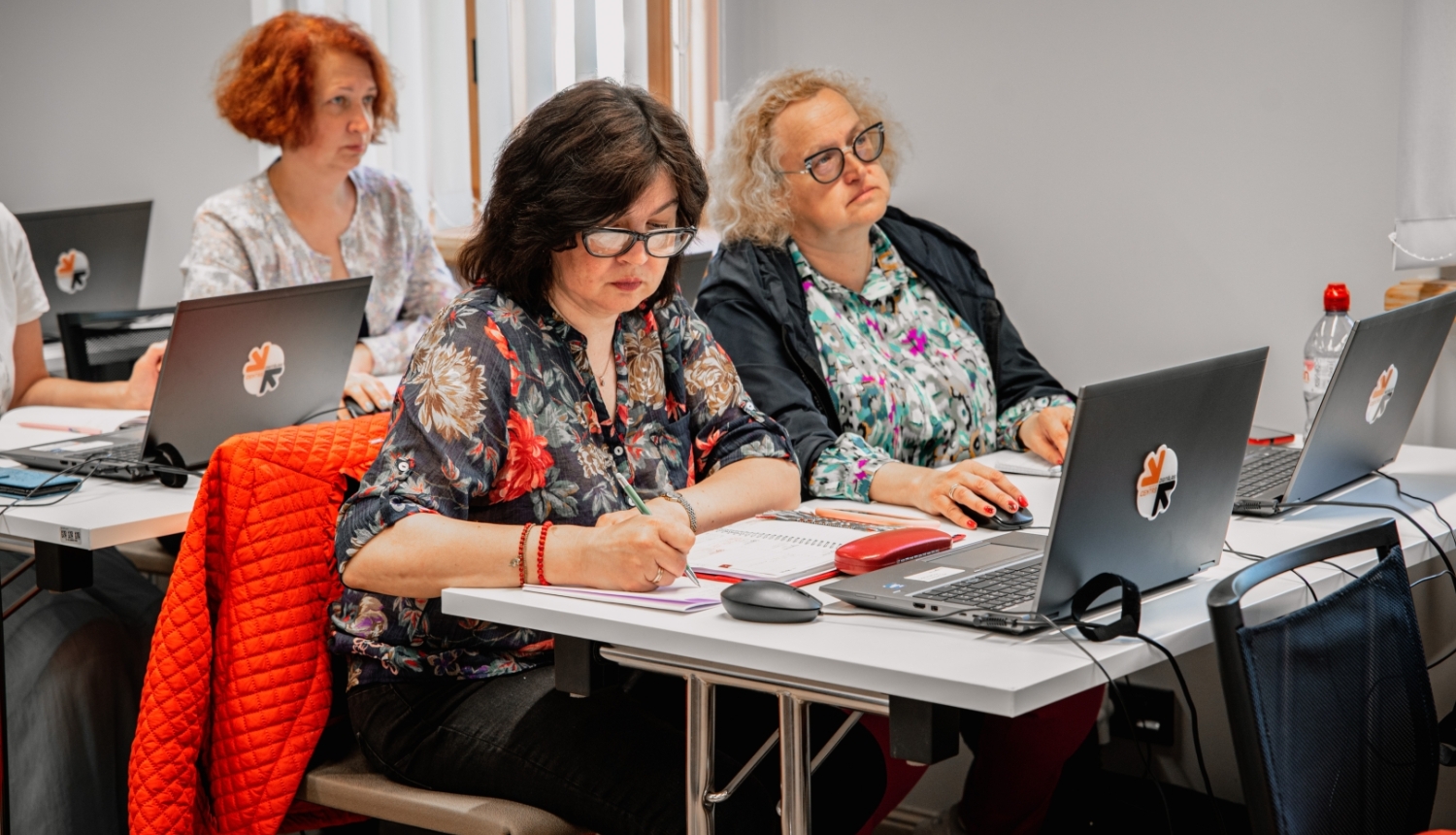 Telpā pie galdiem vairākas sievietes strādā ar klēpjdatoriem un pierakstu kladēm, viena raksta piezīmes, pārējās koncentrējas uz darbu.