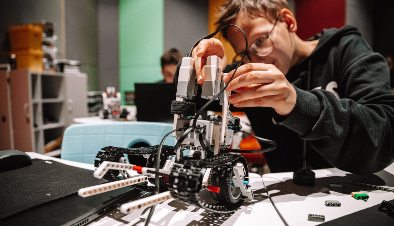 Zēns ar brillēm saliek LEGO robotu, strādājot pie galda tehniski aprīkotā telpā, fonā redzami citi bērni pie datoriem.