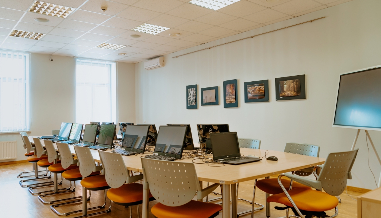 Plaša datoru klase ar portatīvajiem datoriem uz galdiem, krēsliem ar oranžām sēdvietām un vairākām fotogrāfijām uz sienas.