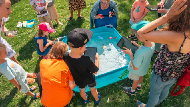 Bērni un pieaugušie brīvā dabā apkārt nelielam baseinam lieto planšetdatorus un viedtālruņus