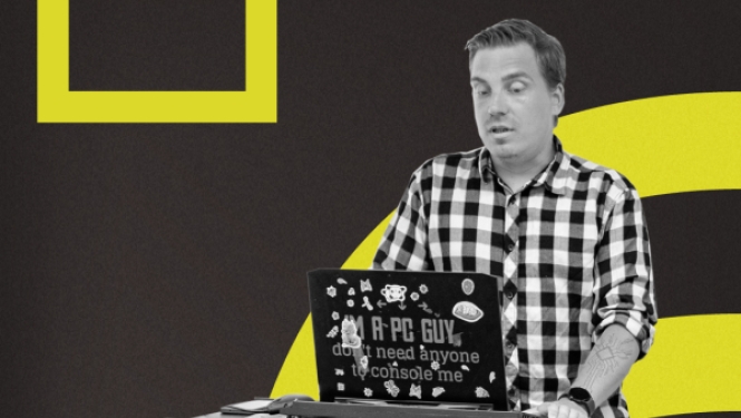 Vīrietis rūtainā kreklā stāv pie augstā galda un strādā ar klēpjdatoru, kura vāks ir noklāts ar uzlīmēm, uz tumša fona ar dzelteniem grafiskiem elementiem.