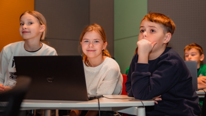 Trīs bērni sēž pie galda ar klēpjdatoru un uzmanīgi klausās nodarbību.
